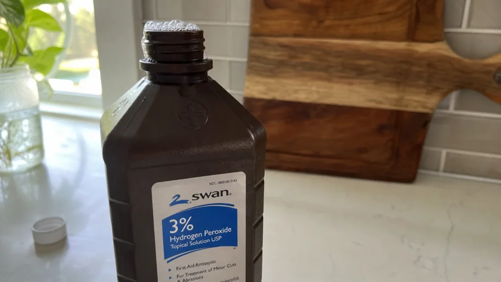 A brown bottle of hydrogen peroxide sits on a white kitchen counter with small bubbles fizzing at the opening.