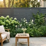 A beautiful backyard with a modern charcoal gray fence, lush green plants, and warm teak wood patio furniture in the golden hour light.