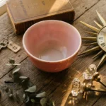 A flat-lay photo of vintage collectibles, including a pink Pyrex bowl, a brass sunburst clock, and crystal doorknobs on a wooden table.