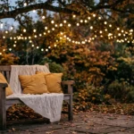 A cozy backyard patio at twilight with string lights glowing above a wooden bench draped with a wool blanket and yellow pillows.