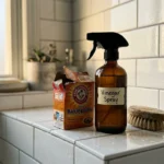 A DIY cleaning setup on a bathroom ledge with a glass vinegar spray bottle, baking soda, and a wooden scrub brush on white tile.