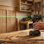 A modern cordless impact driver sits on a wooden workbench with a green laser level casting a alignment line on a wall in the background.