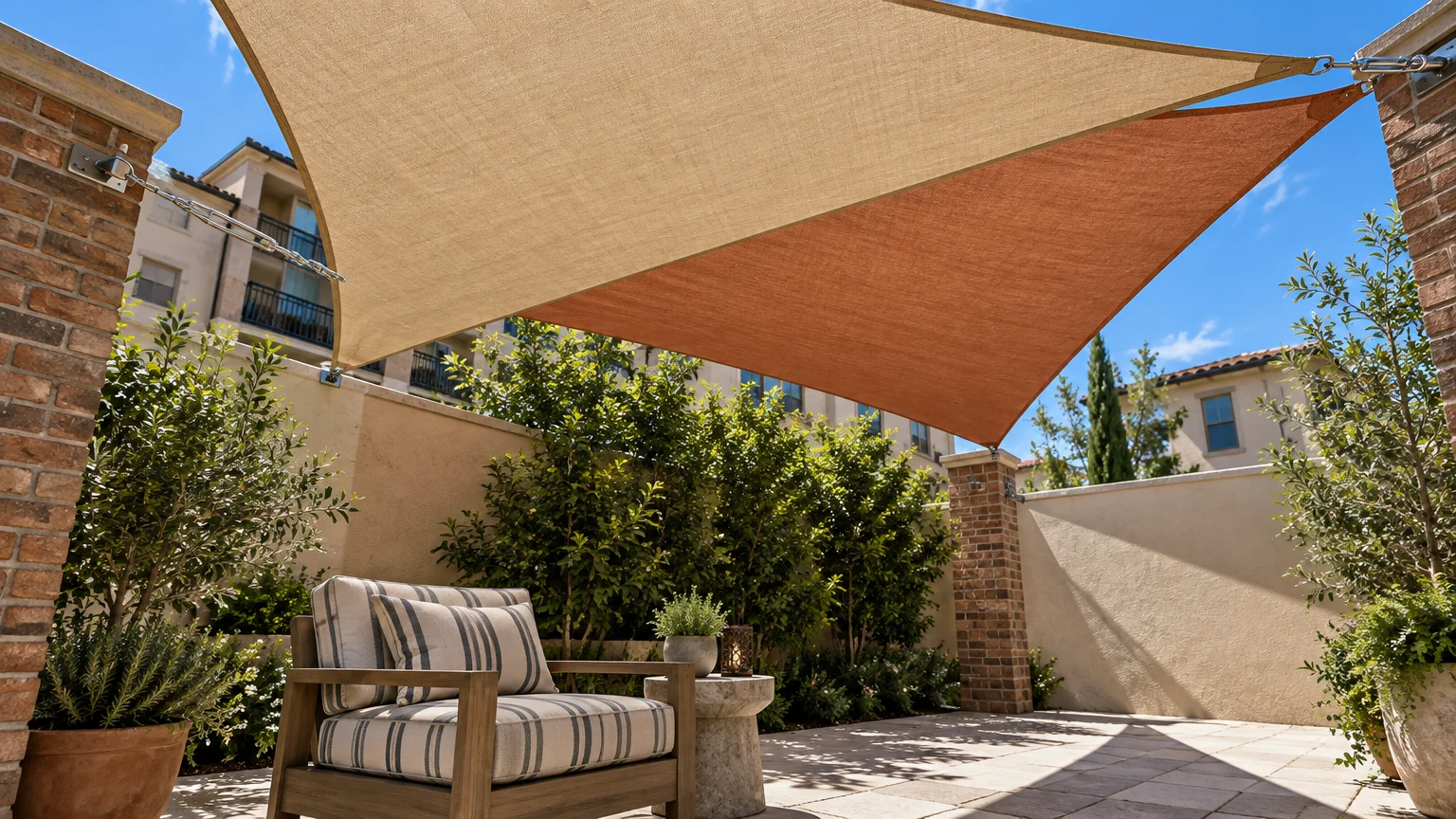 Tan and terracotta shade sails stretched over a stone patio with a striped armchair, plants, and brick pillars.