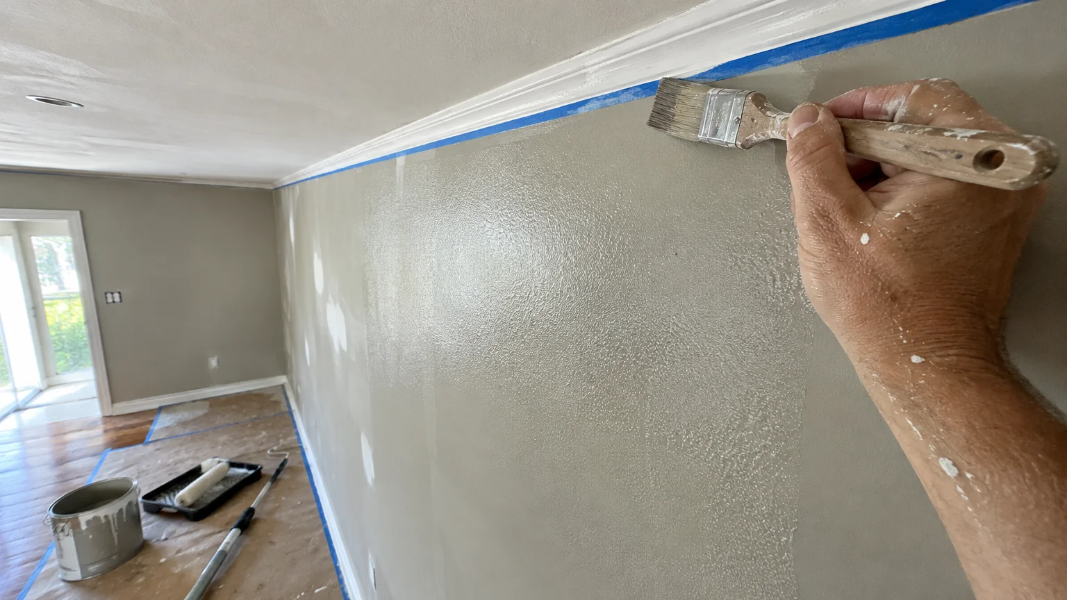 A hand uses a brush to paint a wall edge along blue painter's tape near crown molding in a room undergoing painting.